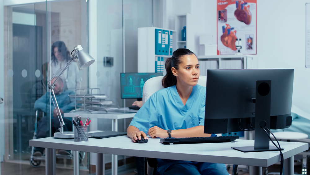 A female NHS staff member works on computer in busy ward